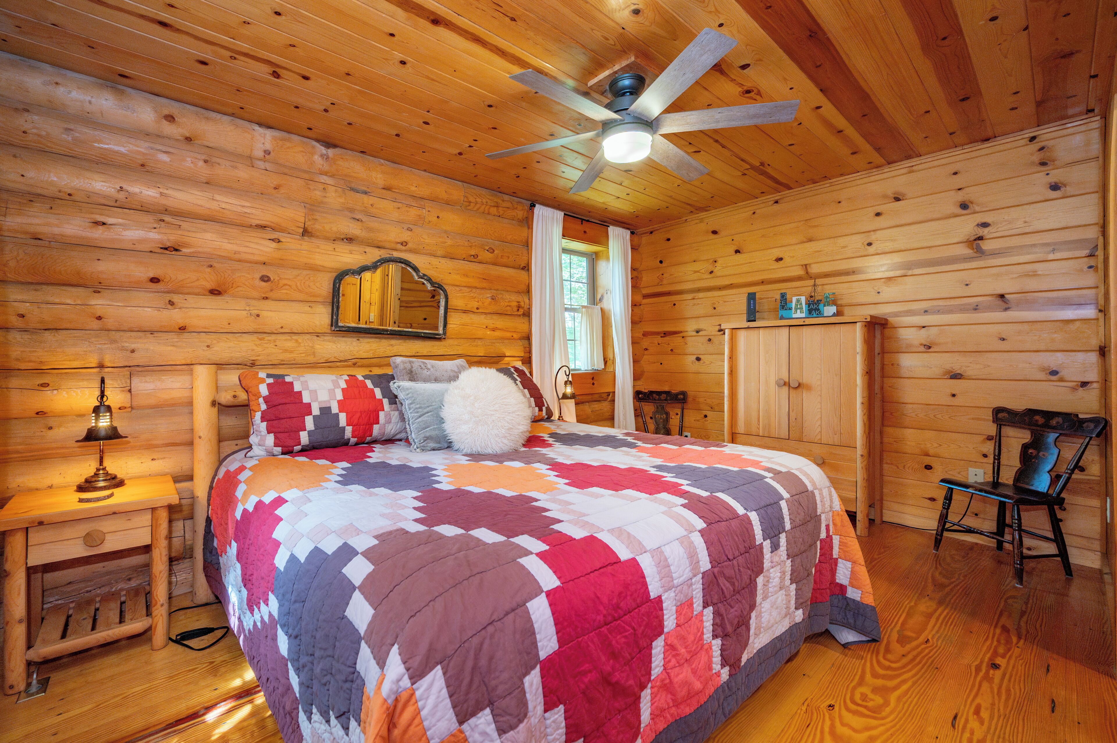 A wooden cabin bedroom with a bed, a nightstand, a chair, a wardrobe, and a ceiling fan.