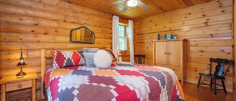A wooden cabin bedroom with a bed, a nightstand, a chair, a wardrobe, and a ceiling fan.