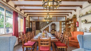 Comedor con una larga mesa de madera dispuesta para una comida, flanqueada por dos sillones azules, una escalera al fondo y un candelabro.