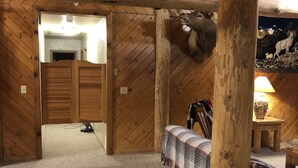 Une pièce rustique avec des poutres en bois, une tête d’animal empaillée accrochée au mur, un lit recouvert d’une couverture à carreaux et une table sur laquelle est posé un vase.