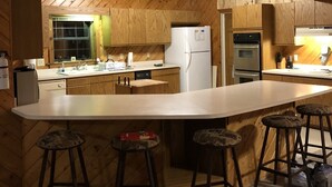 Un intérieur en bois comprenant un espace cuisine, avec un grand îlot central doté de tabourets, un réfrigérateur et une cuisinière.