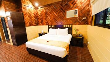 A hotel room with a wooden herringbone-patterned ceiling, a bed with white linens, a nightstand with a lamp, and a window with a view of greenery.