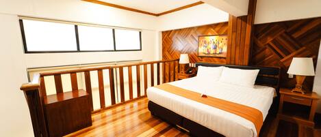 Habitación de hotel con piso de madera, una cama grande con ropa blanca, cabecera de madera, mesita de noche con lámpara y un cuadro en la pared.