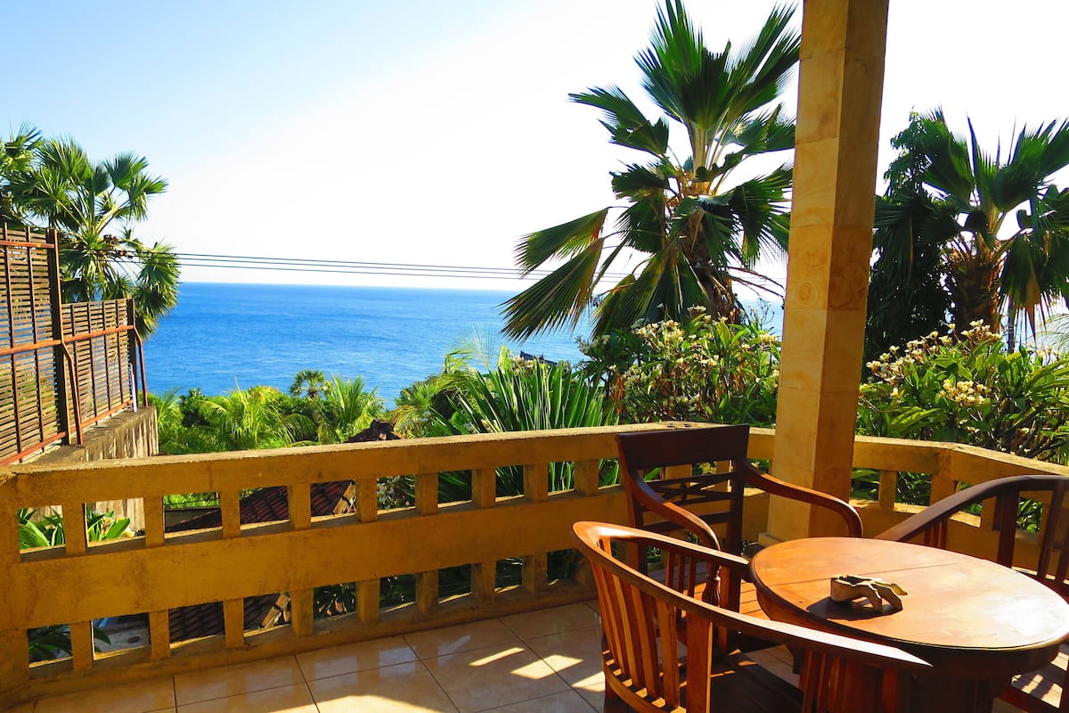 A balcony with a view of the ocean, wooden furniture, and tropical plants.