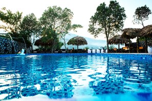 A swimming pool with clear blue water, surrounded by trees and thatched umbrellas. - Ha Giang Moments Homestay - Hostel (Ha Giang 1)
