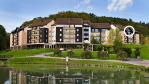 A multi-story residential building with balconies, a reflection in a calm body of water, and a green landscape with trees and grass.