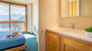 A bedroom with a bed, a window with a view of mountains, a bathroom with a sink and mirror, and wooden cabinets.
