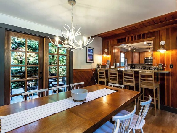 A dining area with a long wooden table, white chairs, a chandelier, and a view through large windows.