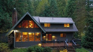 Une cabane à deux étages avec une grande fenêtre à l’étage supérieur, une terrasse et une cheminée.