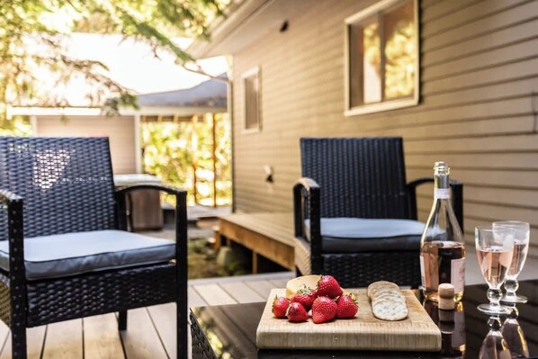 Une terrasse avec des fauteuils en osier, une table en bois sur laquelle se trouve une planche à découper, des fraises, du fromage, une bouteille de vin et des verres.