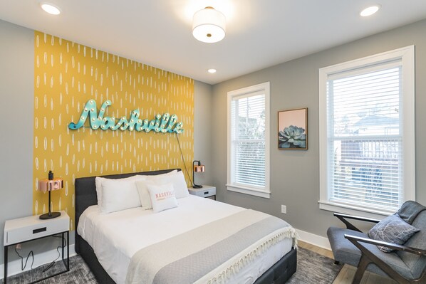 Une chambre à coucher comprenant un lit, des tables de chevet, une chaise, un luminaire fixé au mur et une fenêtre avec des stores.