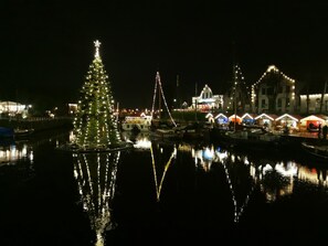 Une scène au bord de l’eau, avec un sapin de Noël et des bâtiments illuminés dont les reflets se mirent dans l’eau.