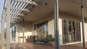 A covered outdoor area with a wooden deck, a seating area with cushions, and a sliding glass door leading to a room.