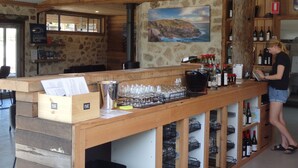 A rustic bar area with wooden walls, a stone wall, and a painting of a lighthouse. There are shelves with bottles and glasses, and a woman is standing at the bar.