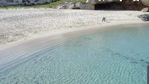 Uma praia calma com uma pessoa caminhando na areia e um afloramento rochoso ao fundo.