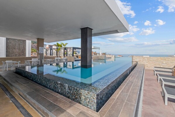 A rooftop infinity pool with a clear view of the ocean, surrounded by lounge chairs and a modern architectural design.