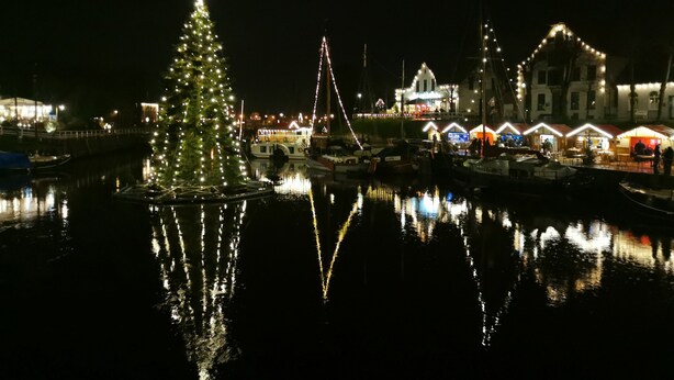 A waterfront scene with a Christmas tree and illuminated buildings reflecting on the water.