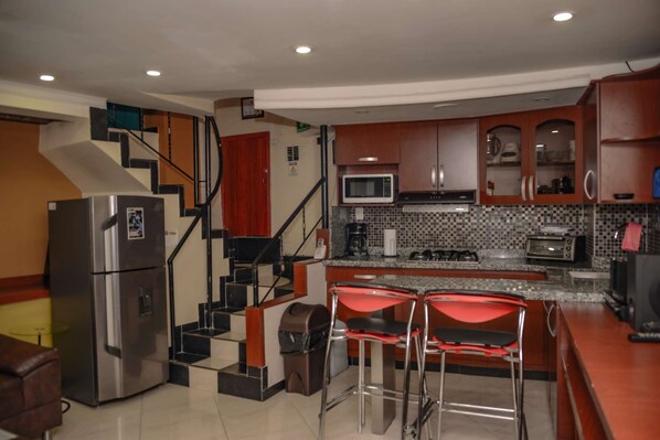 A modern kitchen with stainless steel appliances, a microwave, and a gas stove. There are two red bar stools and a staircase leading to an upper level.