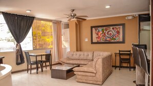Un salon avec un canapé d’angle beige, une table basse, une table à manger avec des chaises, un ventilateur de plafond et un grand tableau abstrait accroché au mur.