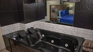 A modern bathroom with a black bathtub, white marble tiles, and a large mirror reflecting a bedroom with a blue bedspread. - King One bedroom AC HOT TUB ELEVATOR 2 Flat screens (Medellin)