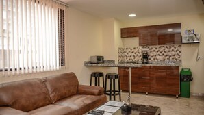 A living room with a brown leather sofa, a coffee table, and a TV. There is a kitchen area with wooden cabinets, a tiled backsplash, and a coffee maker. - King One bedroom AC HOT TUB ELEVATOR 2 Flat screens (Medellin)