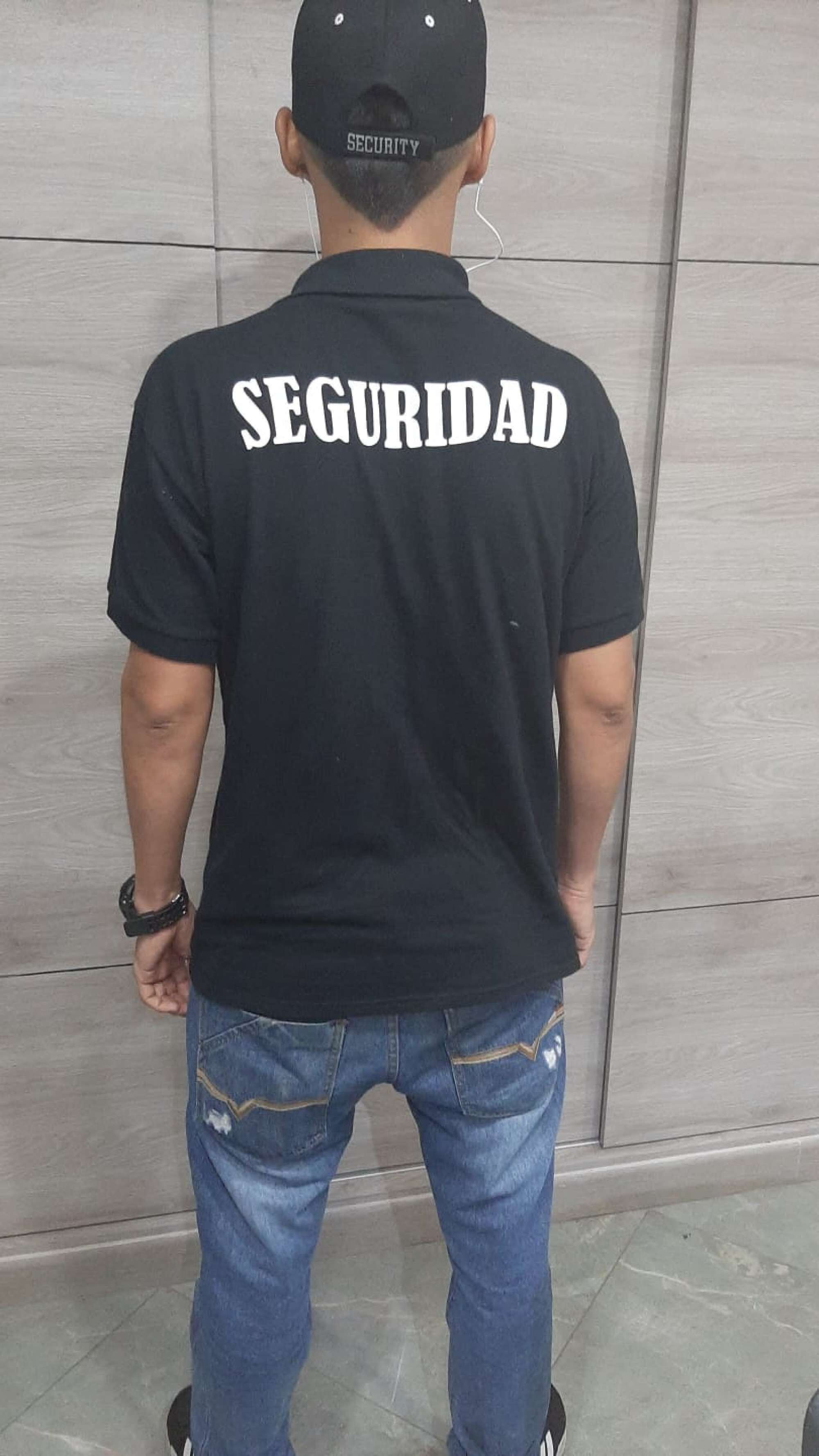 Eine Person mit schwarzem T-Shirt, auf dessen Rückseite „SEGURIDAD“ aufgedruckt ist, steht vor einer gefliesten Wand.