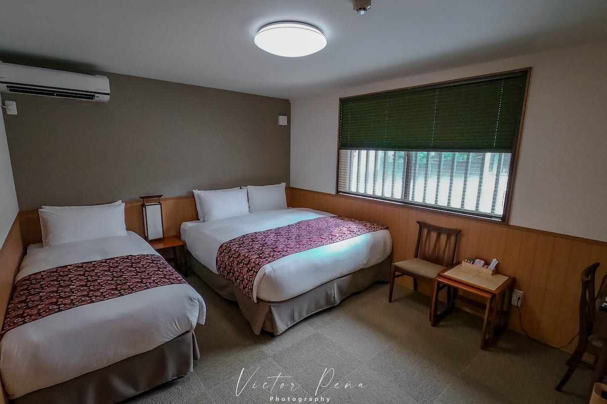 A hotel room with two beds, a small table, a chair, a window with blinds, and an air conditioning unit.