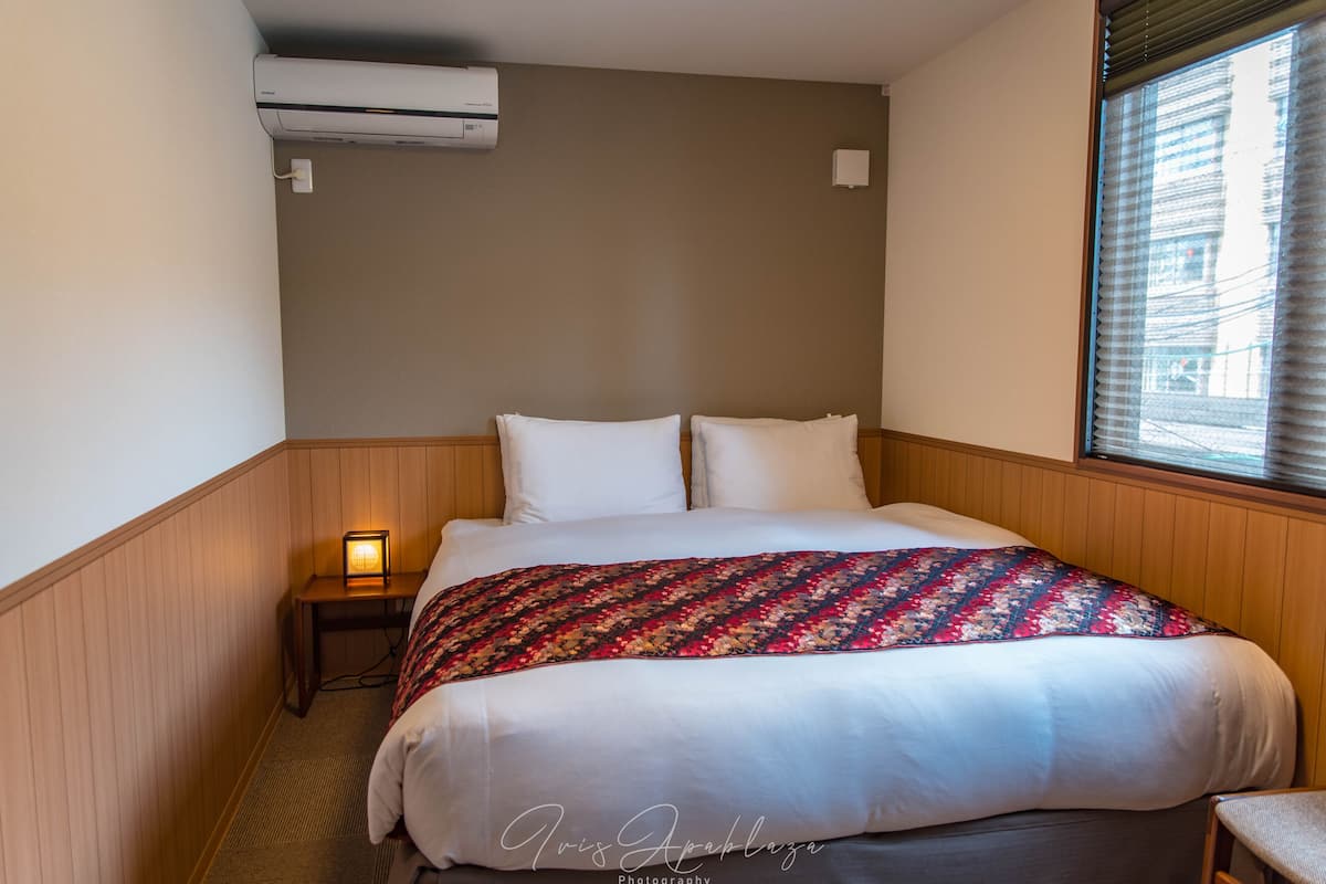 A hotel room with a bed, a nightstand with a lamp, an air conditioner, and a window with blinds.