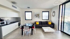 Een modern appartement met keuken, eetruimte en woonkamer. Het heeft een eettafel met stoelen, een bank en een salontafel. Twee ramen bieden uitzicht op de stad.