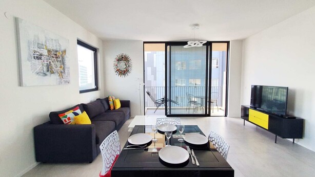 Een moderne woonkamer met een bank, een eettafel voor vier personen, een televisie en een balkon met uitzicht over de stad.