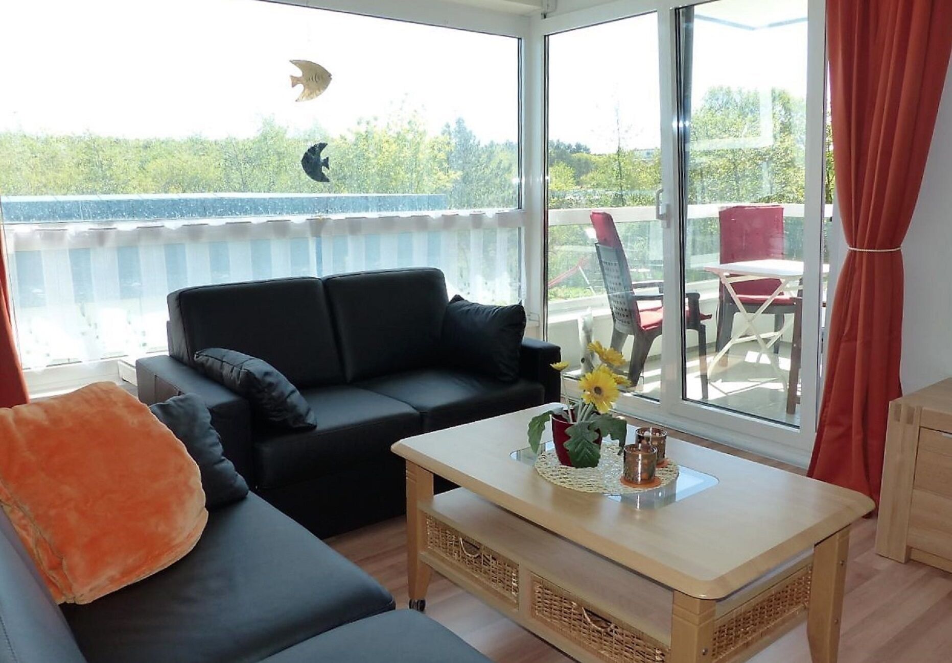 Sala de estar con un sofá negro, una mesa de centro de madera con un florero y vistas a la vegetación a través de amplias ventanas.