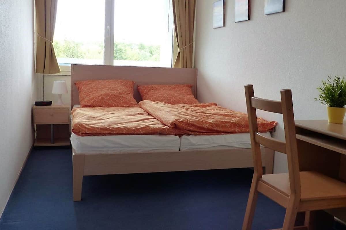 A bedroom with a bed, a desk, a chair, a nightstand, a lamp, and a potted plant.