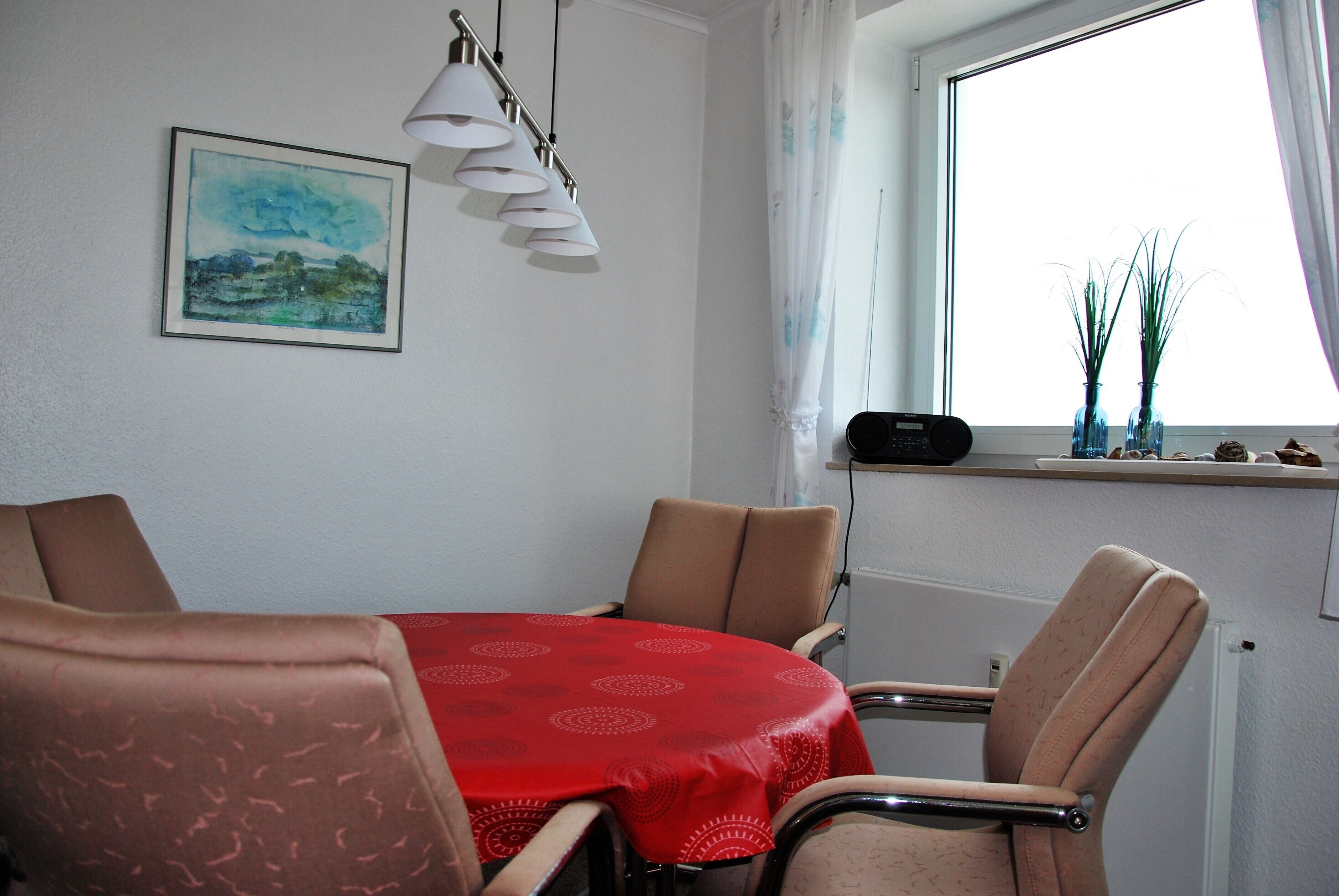 A dining area with a red table, four chairs, a painting, and a window with curtains.