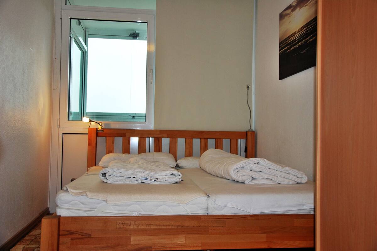 A small, single-bed room with a wooden headboard, a window with a view, and a wall-mounted picture.