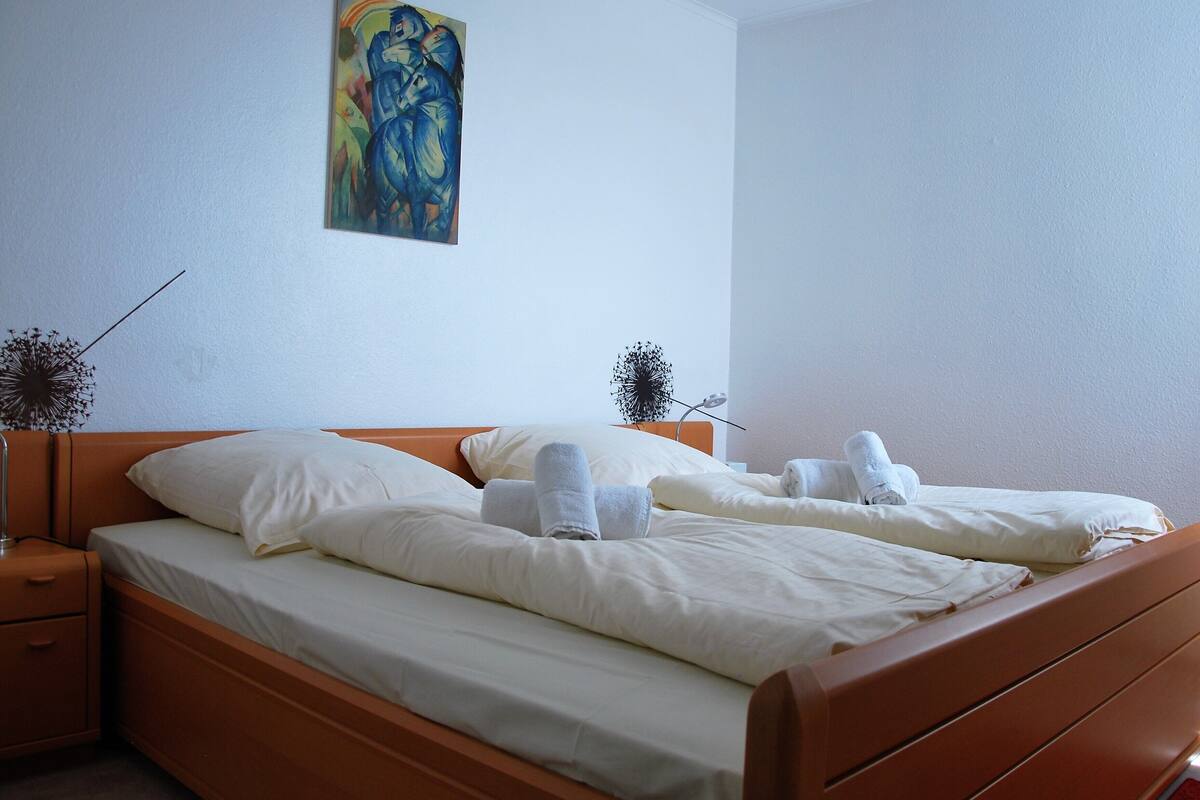 A double bed with white linens and towels, a nightstand, and a painting on the wall.