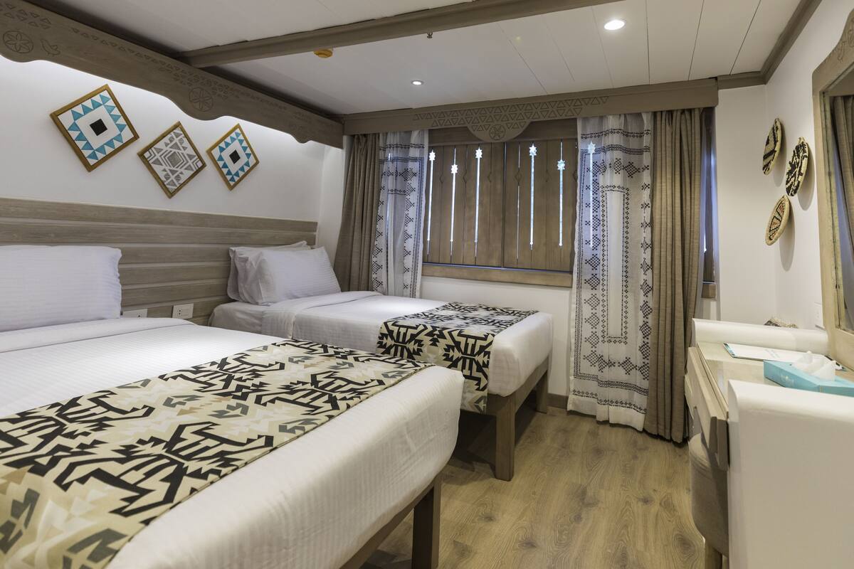 A hotel room with two beds, a wooden headboard, patterned curtains, and a small bedside table.