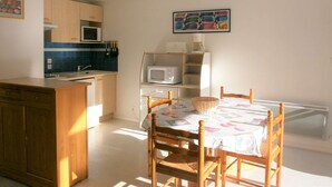 A compact kitchen with a microwave, sink, and wooden cabinets. There is a dining table with chairs and a wall-mounted shelf above it.