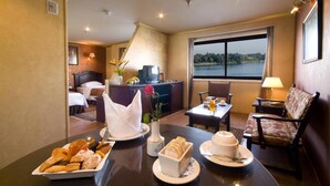 Habitación de hotel con una mesa de comedor dispuesta con pasteles y un jarrón de flores, una cama con televisor, un escritorio con computadora y vista a un lago.