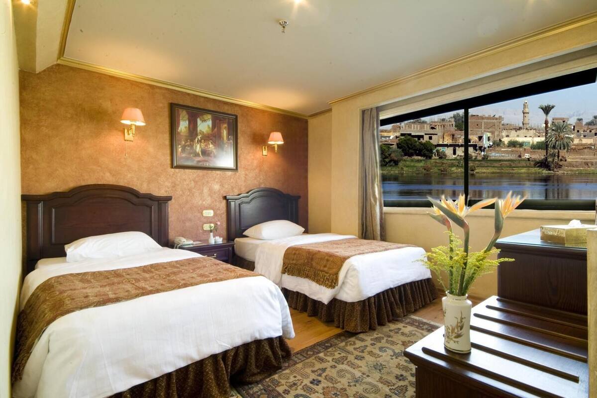 Une chambre d’hôtel avec deux lits, une grande fenêtre donnant sur le paysage urbain et un vase rempli de fleurs placé sur une table de chevet.