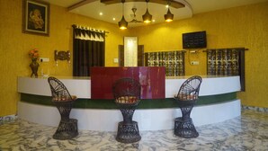 A reception area with a curved counter, three wireframe chairs, a framed picture on the wall, a decorative vase, and a digital display. - Vacation Village Luxury Tent - A Unit Of Braham Vatika Resort (Pushkar)