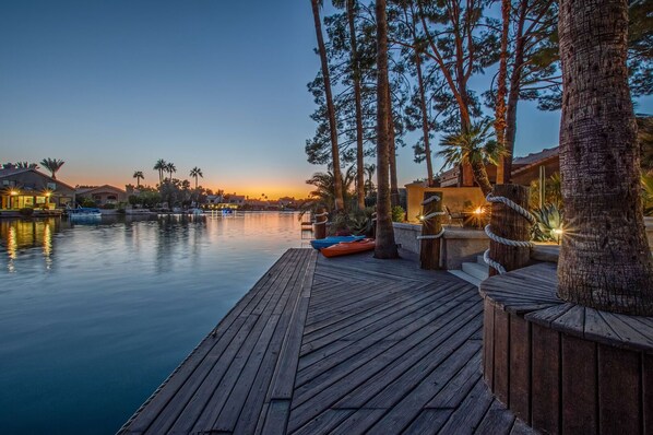 A wooden deck extending over calm water, with a boat and lounge chairs.