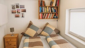 Quarto com cama, mesa de cabeceira, luminária, estante de livros e janela com persianas.