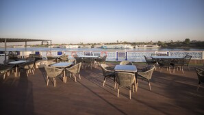 An outdoor seating area with tables and chairs, overlooking a body of water and a distant cityscape. - SUNRISE Semiramis III Cruise (Luxor)