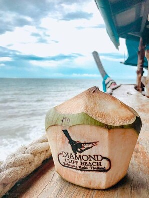 A coconut with a label reading "DIAMOND CLIFF BEACH Koh Lanta Koh Lanta Thailand" on a wooden surface by the sea. - Loft Vintage Chalet Resort (Ko Lanta)