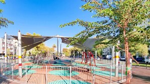 An outdoor playground with swings and slides, surrounded by a metal fence and trees. - Charming 3bdr apt with sea view B5 (Bat Yam)