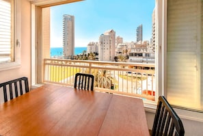 A balcony with a wooden table and chairs, overlooking a cityscape with tall buildings and a clear blue sky. - Charming 3bdr apt with sea view B5 (Bat Yam)