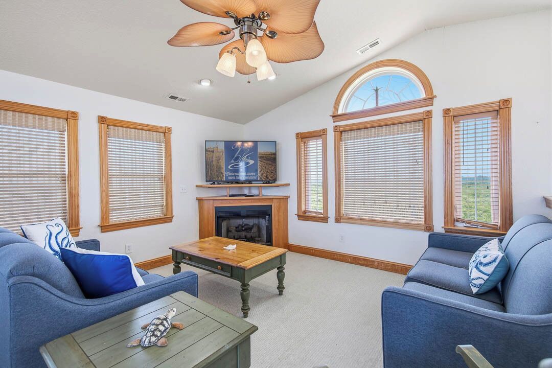 A living room with a fireplace, a ceiling fan, and large windows with blinds.