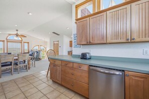 A kitchen with wooden cabinets, a stainless steel dishwasher, and a dining area with a table and chairs. - The After Glow | 940 ft from the beach | Private Pool, Hot Tub (Nags Head)