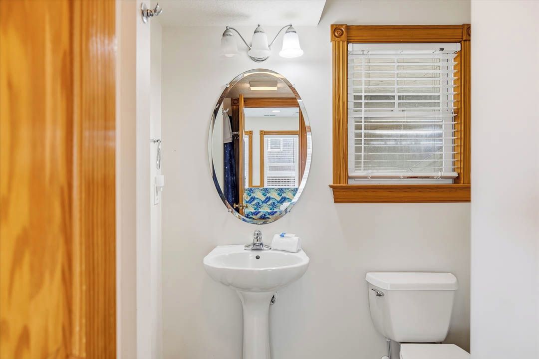 Un baño con lavamanos blanco, espejo, inodoro y ventana con persianas.