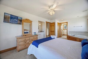 Een hotelkamer met een bed, een houten dressoir, een spiegel, een plafondventilator, een badkamer met een bad en een ingelijst schilderij aan de muur.